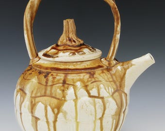 May include: A ceramic teapot with a rounded body, featuring a cream-colored base glaze with brown drip patterns. It has a high, arched handle, a short spout, and a lid topped with a textured, stem-like knob. The surface has a glossy finish.