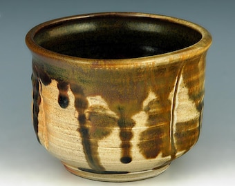 May include: A ceramic bowl with a rounded shape and a wide opening. The exterior features a cream-colored base with dark brown and olive green glaze drips running vertically from the rim. The interior is finished in a deep, dark brown glaze.