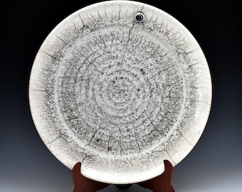 May include: A round ceramic plate featuring a white base with a prominent black crackle glaze pattern. The surface displays a dense, dark web of fine lines radiating from the center, with a small circular metallic accent near the top rim.