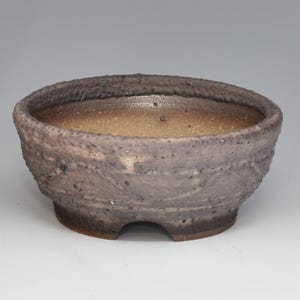 May include: A round, low-profile ceramic bonsai pot with a textured, dark grey and brown exterior. The interior features a smooth, light tan glazed finish. The base has a small, arched cutout, and the rim is thick with a rough, uneven surface texture.