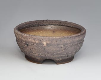 May include: A round, low-profile ceramic bonsai pot with a textured, dark grey and brown exterior. The interior features a smooth, light tan glazed finish. The base has a small, arched cutout, and the rim is thick with a rough, uneven surface texture.