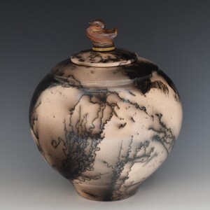 May include: A ceramic jar with a lid, featuring a duck-shaped handle. The jar has a round shape with a light beige base and black, lightning-like patterns. The lid is beige with black markings and a gold-colored band.