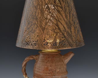 May include: A handcrafted ceramic lamp with a teapot-shaped base in shades of brown and cream. The lamp features a textured, patterned lampshade in brown, topped with a dark wooden finial. The lamp sits on a dark brown wooden base.