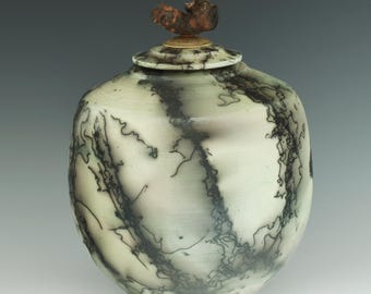 May include: A ceramic vessel with a smooth, off-white surface featuring dark, intricate, web-like black lines. The vessel has a matching lid topped with a small, textured, brown wooden knob. The overall shape is rounded with a tapered base.