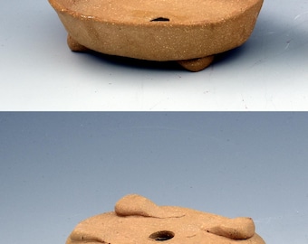 May include: A shallow, oval-shaped terracotta planter with a textured, earthy brown surface. The vessel features a wide, open top, a single drainage hole at the base, and is supported by three small, sturdy feet integrated into the bottom design.
