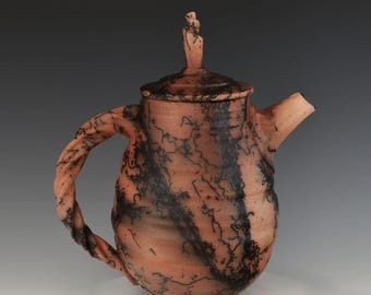 May include: A ceramic teapot with a terracotta-colored body featuring black horsehair-style markings. The teapot has a twisted handle, a lid with a small finial, and a short spout. The surface has a matte finish with horizontal ridges.