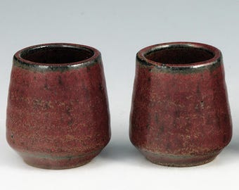 May include: Two handmade, red ceramic cups with a textured surface and a dark rim. The cups have a rounded shape and are displayed against a white background. The cups are likely for drinking.