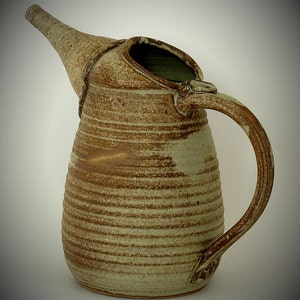 May include: A handmade ceramic pitcher, approximately 9.5 inches high and 11 inches wide. The pitcher has a unique spout and handle design. It features a textured, earthy color palette of brown and beige, with horizontal striped detailing.