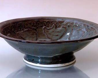 May include: A handmade ceramic bowl with a wide, shallow design. The bowl features a dark teal glaze with a textured, brown rim and interior detailing. It sits on a pedestal base with a white accent.