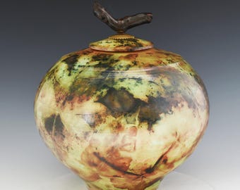 May include: A round, lidded vessel with a smooth, bulbous body featuring a mottled pattern of green, yellow, and brown tones. The lid is topped with a dark, textured, branch-like handle that extends upward, creating a natural, organic aesthetic.