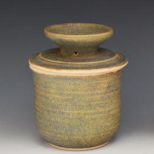 May include: A cylindrical ceramic butter keeper with a mottled olive green and brown glaze. The vessel features a wide, flared top opening with a small circular hole on the side of the neck and a textured, horizontal ridged surface throughout.