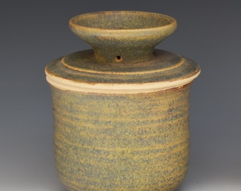 May include: A cylindrical ceramic butter keeper with a mottled olive green and brown glaze. The vessel features a wide, flared top opening with a small circular hole on the side of the neck and a textured, horizontal ridged surface throughout.