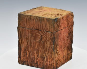 May include: A square, wooden-textured box with a lid. The box is primarily brown with darker accents, and features a carved seashell design on the front. The box has a rustic, handcrafted appearance.