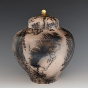 May include: A ceramic urn with a lid, featuring a light beige base with intricate black, lightning-like patterns. The lid is topped with a small, gold-colored knob. The urn's design has a natural, organic aesthetic.