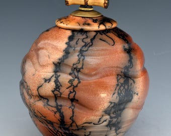 May include: A handcrafted, round ceramic jar with a lid. The jar is primarily reddish-brown with black, lightning-like patterns. The lid has a bamboo handle and a brass base. The jar's shape is slightly irregular, adding to its unique appeal.