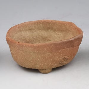 May include: A small, round, terracotta-colored ceramic bowl with a textured, speckled surface. The bowl features a thick, uneven rim, a deep interior, and is supported by three small, rounded feet at the base. A small, circular stamp is visible.