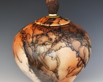 May include: A handcrafted ceramic urn with a unique, organic design. The urn features a rounded body with a lid and a textured, dark brown knob. The surface is a blend of tan and orange with intricate black patterns.