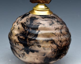 May include: A handcrafted ceramic vessel with a bulbous shape, featuring a brown and black abstract design. The vessel has a gold-colored metal collar and a wooden stopper. The overall aesthetic is rustic and artistic.