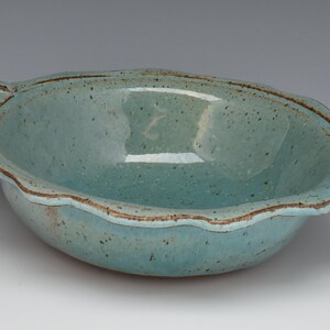 May include: A handmade, turquoise ceramic bowl with a scalloped rim and two decorative handles. The bowl has a speckled texture and a brown rim. This unique piece is perfect for serving or display.