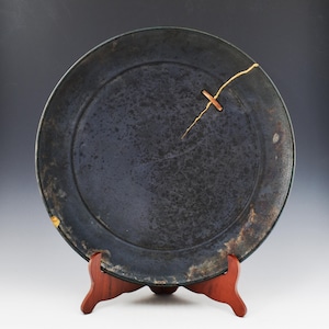 May include: A dark blue ceramic plate with a visible crack repaired with gold-colored lacquer. The plate is round and sits on a small, reddish-brown wooden stand. The plate has a textured surface and a slightly uneven rim.