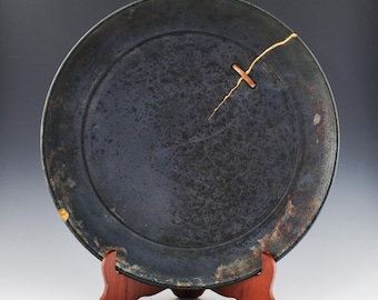 May include: A dark blue ceramic plate with a visible crack repaired with gold-colored lacquer. The plate is round and sits on a small, reddish-brown wooden stand. The plate has a textured surface and a slightly uneven rim.