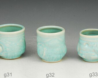 May include: Five small, handmade ceramic cups with a light turquoise glaze. Each cup is approximately 2 inches tall and wide. The cups have a textured surface and a contrasting darker turquoise interior.