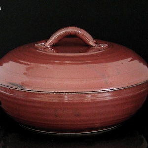 May include: A maroon ceramic casserole dish with a lid and handles. The dish is approximately 5 inches high and 10 inches wide. The lid has a curved handle. The dish is on a black background.