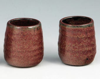May include: Two handmade, textured ceramic cups. The cups are a deep red with a speckled pattern and a dark rim. The cups have a rustic, organic shape, perfect for enjoying a warm beverage.