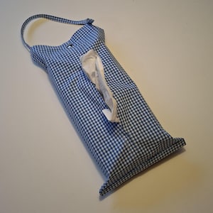 Pode incluir: Um porta-lenços retangular de tecido com um padrão de xadrez vichy azul e branco. O suporte inclui uma alça superior para pendurar e uma abertura vertical central, com um lenço de papel branco a sobressair do centro da bolsa.