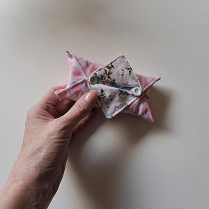 Könnte beinhalten: Ein kleines Stoffetui mit einer rosa-weiß karierten Außenseite und einer floral gemusterten Innenklappe. Das Etui wird mit zwei weißen Knöpfen verschlossen und bildet eine gefaltete Umschlagform, die einen kleinen Gegenstand in der Öffnung hält.