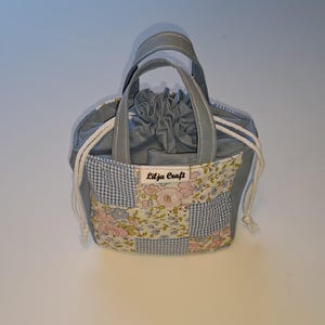 Op de afbeelding: Een kleine stoffen tas met trekkoord en twee handvatten, voorzien van een patchworkontwerp van blauwe gingham en bloemenpatronen in roze, blauw en groen. Een wit label met de tekst Lilja Craft is op de voorzijde genaaid met witte koorden.