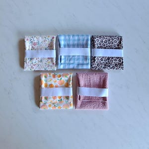 May include: Five folded fabric items with white elastic bands. The fabrics feature various patterns: floral, blue gingham, leopard print, orange fruit, and red and white check. The items are arranged on a white surface.