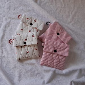 Peut inclure: Quatre pochettes matelassées. Deux sont blanches avec un motif de cerises, une fermeture à bouton noir et un anneau rouge. Les deux autres sont à rayures roses et blanches avec une fermeture à bouton noir.