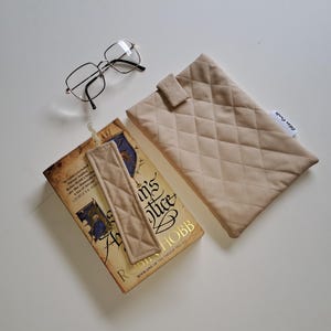 Könnte beinhalten: Ein beige gesteppter Buchumschlag, ein Lesezeichen und eine Brille liegen auf einer weißen Oberfläche. Das Buch trägt den Titel "Assassin's Apprentice" von Robin Hobb. Lesezeichen und Hülle haben ein Rautenmuster.