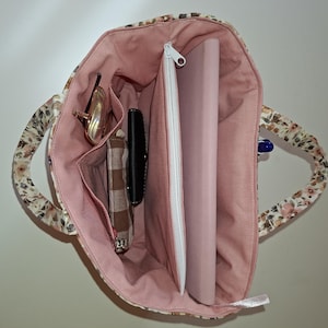 Può includere: Una borsa in tessuto con esterno a motivi floreali e fodera interna color rosa polvere. La borsa contiene uno scomparto con cerniera, una pochette a quadri marroni e bianchi, un portafoglio nero, un paio di occhiali e un tablet rosa.