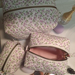 May include: Three quilted cosmetic bags featuring a white background with a small purple floral and green leaf pattern. Each bag has a metallic zip closure. One bag is open, revealing a pink and white gingham interior lining.