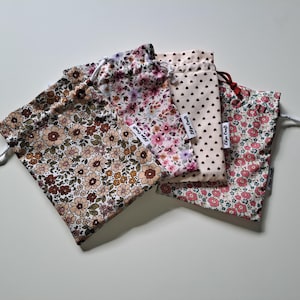 Pode incluir: Quatro bolsas retangulares de tecido com cordão, com vários padrões, incluindo flores castanhas e pêssego, flores cor-de-rosa e roxas, creme com corações pretos e um padrão floral vermelho e verde. Cada bolsa tem uma pequena etiqueta branca Lilja Craft.