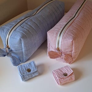 Op de afbeelding: Twee rechthoekige gewatteerde etuis met lichtblauw en lichtroze gingham-ruitpatronen, elk voorzien van een witte rits met een strikvormige trekker. Bij elk etui hoort een klein, bijpassend vierkant tasje met een zilverkleurige drukknoopsluiting.