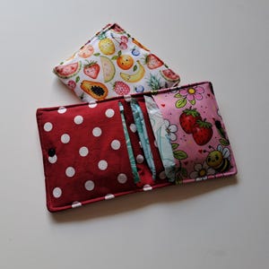 May include: A red polka dot diaper clutch with a snap closure, open to reveal diapers. A matching fruit-patterned changing pad is on top. The clutch has a pink strawberry and bee pattern on the inside.