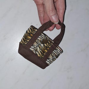 Op de afbeelding: Een miniatuur draagtas met een tijgerstrepenpatroon in zwart, oranje en wit. De tas heeft een effen bruine stoffen rand langs de onderkant en de handvatten, met een enkele ronde bruine drukknoopsluiting in het midden van het voorpaneel.