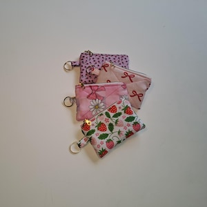 May include: Four small, quilted fabric coin purses in various patterns. Designs include a purple polka dot, pink bows, a daisy, and strawberries. Each purse has a gold zipper and a metal key ring.