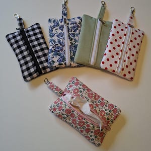 Puede incluir: Cinco estuches rectangulares de tela con cremalleras y cierres de mosquetón de metal. Cuatro estuches están en posición vertical con varios patrones, incluyendo cuadros vichy blanco y negro, flores azules, verde salvia liso y blanco con lunares rojos.
