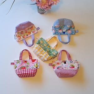 以下が含まれることがあります： イチゴ、レモン、花柄など、さまざまな模様の小さな手作りの布製トートバッグが5つ。各バッグには、お揃いのハンドルとギンガムチェックまたは模様入りのベースが付いています。バッグは白い表面に配置されています。