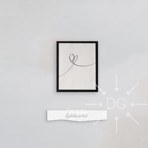 May include: A framed print with a minimalist heart line drawing in a black frame on a white wall. Below the frame is a white paper with the word "lighthearted" printed on it. The artwork is simple and elegant.