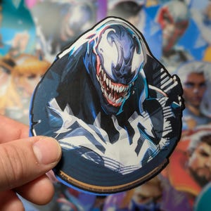 Puede incluir: Una pegatina redonda con un primer plano de Venom, un personaje de superh&eacute;roe ficticio de Marvel Comics. Venom est&aacute; representado con un traje blanco y negro, una boca grande con dientes afilados y una expresi&oacute;n amenazante. La pegatina tiene un fondo azul con un borde dorado.