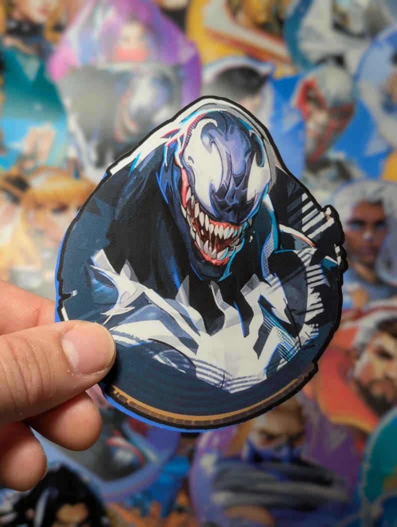 Puede incluir: Una pegatina redonda con un primer plano de Venom, un personaje de superh&eacute;roe ficticio de Marvel Comics. Venom est&aacute; representado con un traje blanco y negro, una boca grande con dientes afilados y una expresi&oacute;n amenazante. La pegatina tiene un fondo azul con un borde dorado.