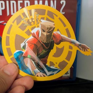 Puede incluir: Una pegatina circular amarilla con un personaje de dibujos animados en rojo y marr&oacute;n que lleva una m&aacute;scara de bolsa de papel marr&oacute;n. El personaje sostiene un rayo amarillo. La pegatina tiene el texto "Marvel's Spider-Man 2" en el fondo.