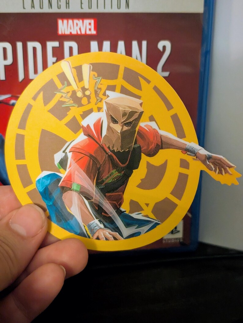 Puede incluir: Una pegatina circular amarilla con un personaje de dibujos animados en rojo y marr&oacute;n que lleva una m&aacute;scara de bolsa de papel marr&oacute;n. El personaje sostiene un rayo amarillo. La pegatina tiene el texto "Marvel's Spider-Man 2" en el fondo.