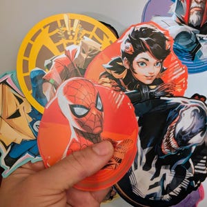 Puede incluir: Una colecci&oacute;n de pegatinas redondas con varios personajes, entre ellos Spider-Man, Venom y un personaje femenino con ojos azules. Las pegatinas est&aacute;n impresas sobre un fondo blanco y tienen un acabado brillante.