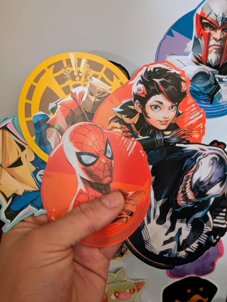 Puede incluir: Una colecci&oacute;n de pegatinas redondas con varios personajes, entre ellos Spider-Man, Venom y un personaje femenino con ojos azules. Las pegatinas est&aacute;n impresas sobre un fondo blanco y tienen un acabado brillante.